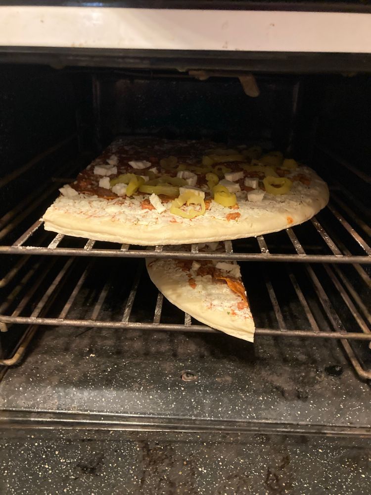 The speckled enamel interior of an oven containing an uncooked pizza. About three quarters of the pizza (width-wise, at least, which is the important dimension here) occupies nearly all of the top rack. The remaining section sits on the second rack beneath it. 
If it matters to anyone, the visible toppings are banana pepper rings and torn-up bits of fresh mozzarella. 