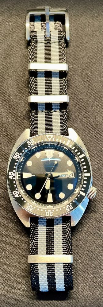 an analogue wrist watch, silver case, black and white bezel, black and white dial, grey and black NATO strap.