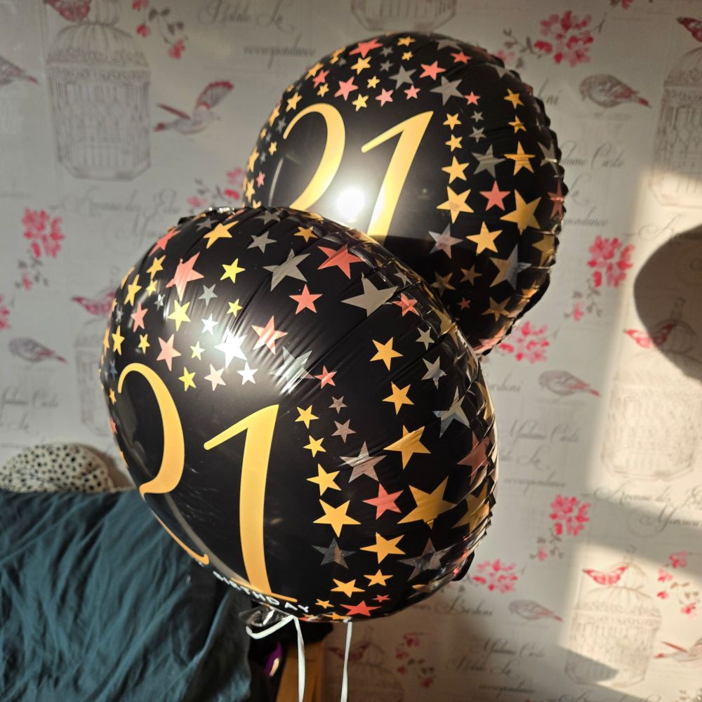 2 balloons with gold, bronze, and silver stars on and large number '21's on in front of some white with pink detailing wallpaper and a green pillow to the lower left.