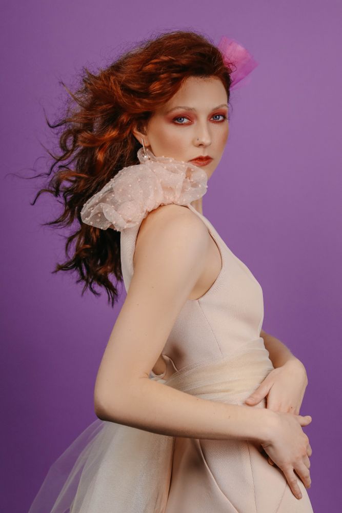 redheaded curly hair model. wearing tan/pink dress with fluffy sleeves and bustle. super dramatic makeup. bright red lip and bright red smokey eye. purple background. 