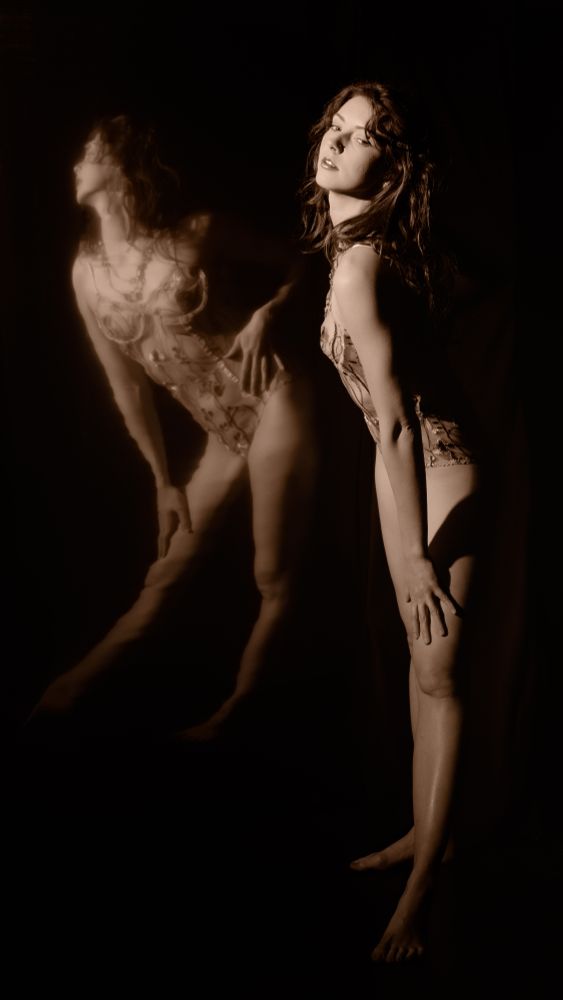sepia toned photo. woman standing in front of mirror with lace floral lingerie. curly hair. bent over touching knee.