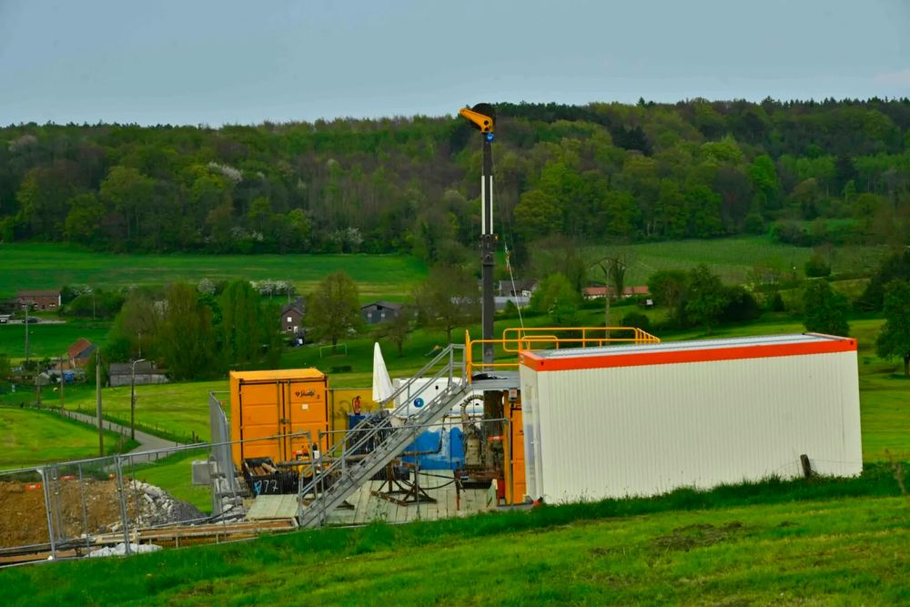 Drilling site in a rural landscape in Sint Pietersvoeren, 2024. The setup includes a tall drilling rig, orange and white machinery containers, and a metal staircase structure. In the background, green fields, scattered houses, and a dense forest stretch across the horizon under a cloudy sky.