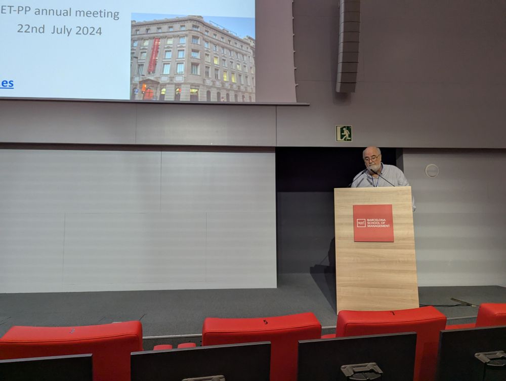 A man standing at a podium inside an auditorium, delivering a presentation during the ET-PP Annual Meeting at the Barcelona School of Management. A slide projected behind him shows the event title “ET-PP annual meeting 22nd July 2024” and a photo of a building.
