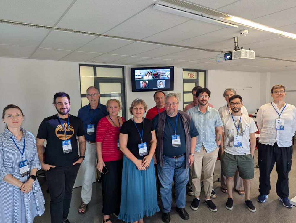 A group of people posing indoors for a group photo during the ET-PP Annual Meeting. They are wearing blue lanyards with name badges, and a video call screen in the background shows additional participants joining remotely.