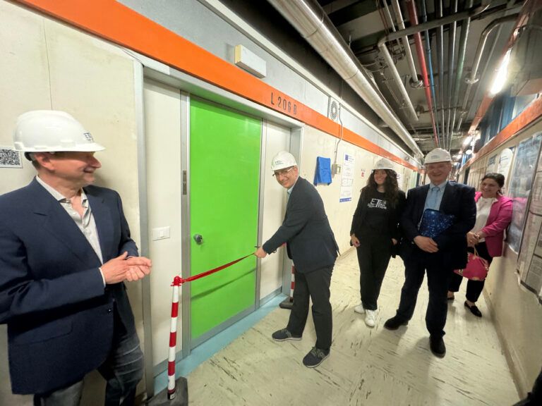 A ribbon-cutting ceremony at the GALILEO laboratory. A group of people in safety helmets stand in an underground corridor. One man, smiling, cuts a red ribbon in front of a bright green door marked L2060. Others, including scientists and officials, look on with interest.