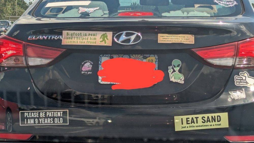 Car with bumper stickers that say "bigfoot is real and I helped him commit tax fraud", "honk if you'd rather be watching the 1999 cinematic masterpiece the mummy starring Brendan Fraser and Rachel Weisz", "please be patient I am 9 years old", "I eat sand just a little some times as a treat"