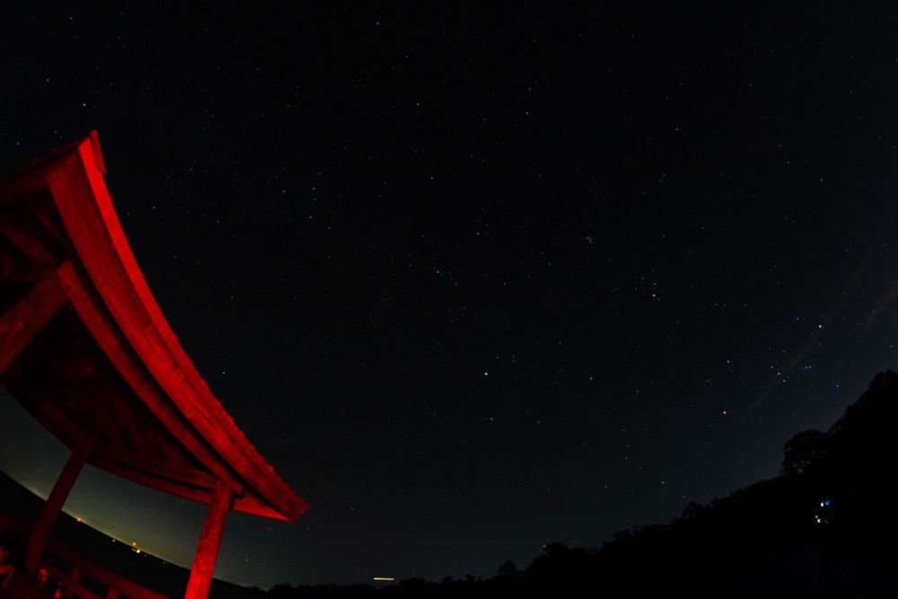 landscape fisheye photo of the night sky aimed somewhat between the milky way and taurus/orion. the horizon bends around the lower edge of the photo. the center and right half of the bottom has a tree line silhouette, the left has some lights from a distant city. near the camera is a square wooden roofed area on the pier arcing up the left side of the image in stark ominous red lighting.