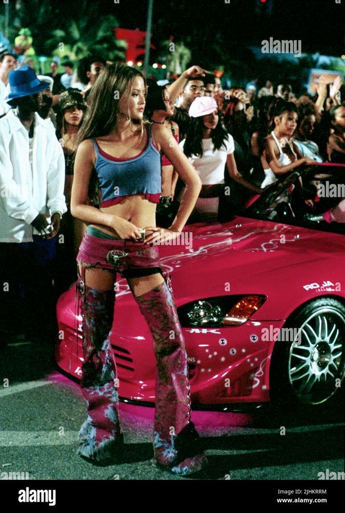 devon aoki as suki in 2 fast 2 furious wearing layered cami crop top tanks and pink denim shorts above distressed pink jean legs attached with suspenders. she is standing in front of a car. very swag