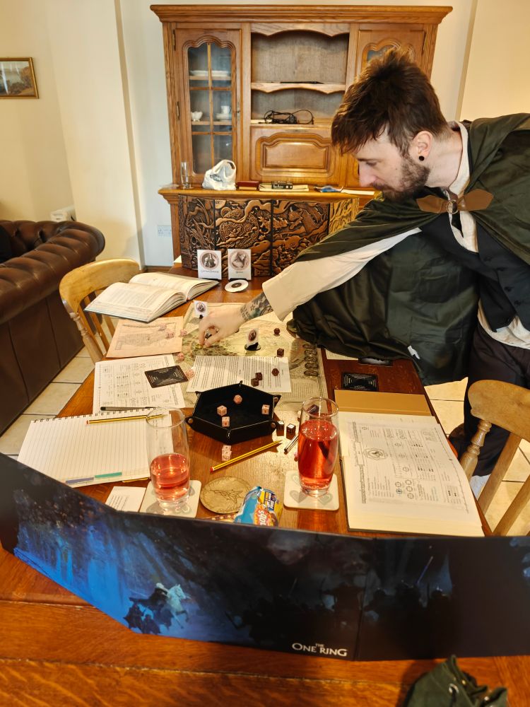 A roleplaying table set up with dice and books. A white guy dressed as a hobbit picks up a die