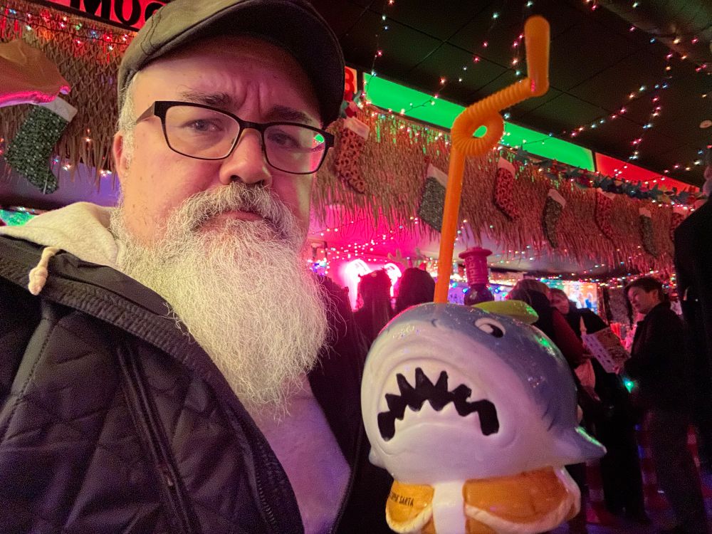 A bearded man, possibly a wizard, in a holiday themed bar drinking from a shark-themed mug. 