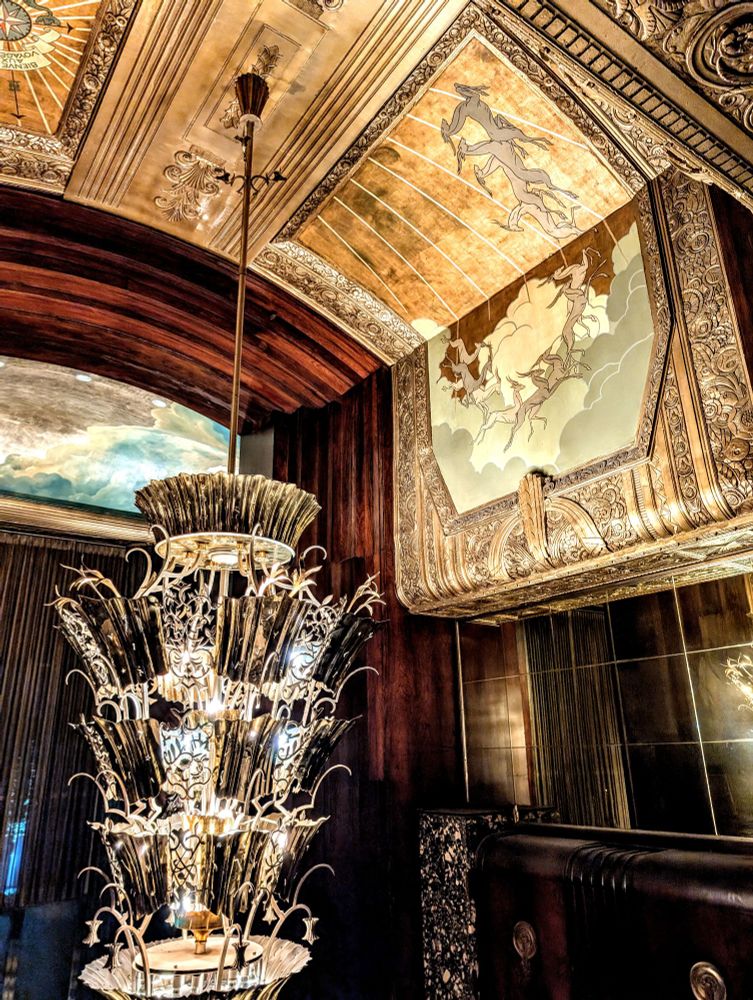 An art Deco chandelier hanging from a ceiling with embellishments, deep wood panels and paintings of flying possibly antelope