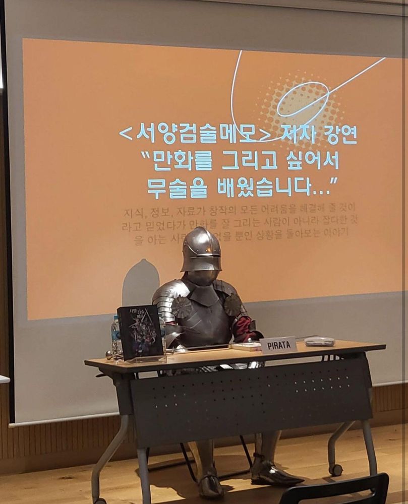 A conference speaker in knight atire seated behind a desk with a slide in Korean behind them.