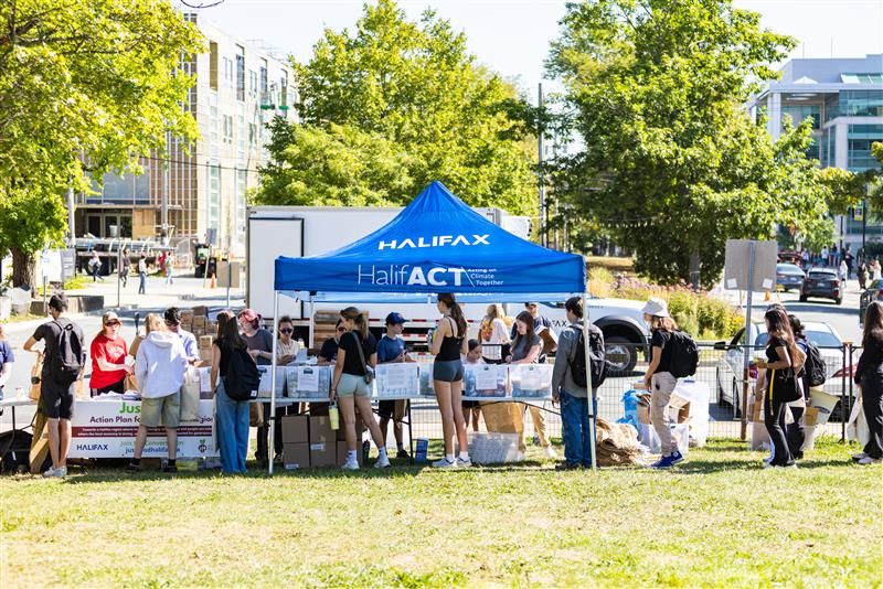 A line of people waiting to build their fresh food stormkits by a row of foldable tables, a blue sun cover that reads "HalifACT" on Dalhousie campus.