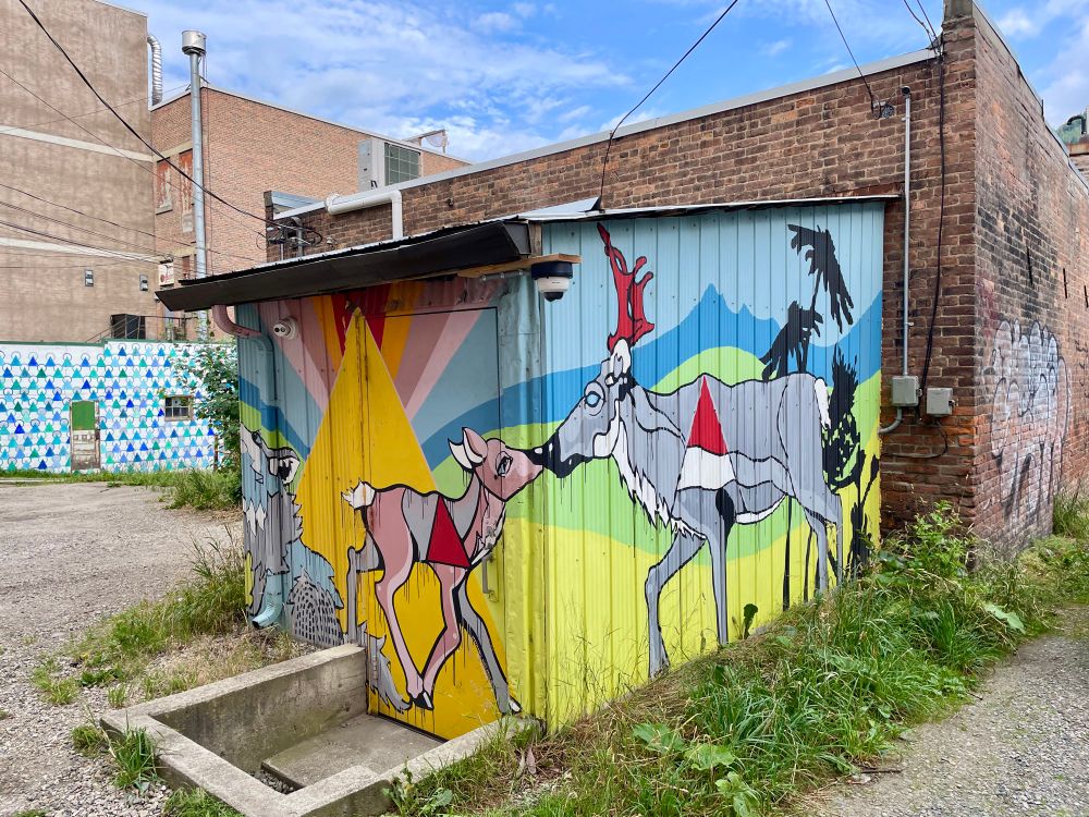 Brightly colored kaleidoscopic alley painting of a small moose or caribou calf touching noses with an adult of the same species. Where their noses touch is the corner of the building. Yellow, blue and green and the predominant colors with pops or red and other colors.