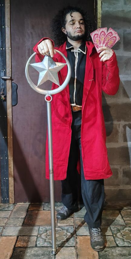 PESSOA VESTIDA DE CALÇA PRETA, BOTAS, SOBRETUDO VERMELHO, UMA CAMISETA COM AS BORDAS BRANCAS POR BAIXO DO SOBRETUDO, COM A MÃO APOIADA EM UM BÁCULO COM UMA ESTRELA DENTRO DE UM CIRCULO, SEGURANDO 4 CARTAS SAKURA;