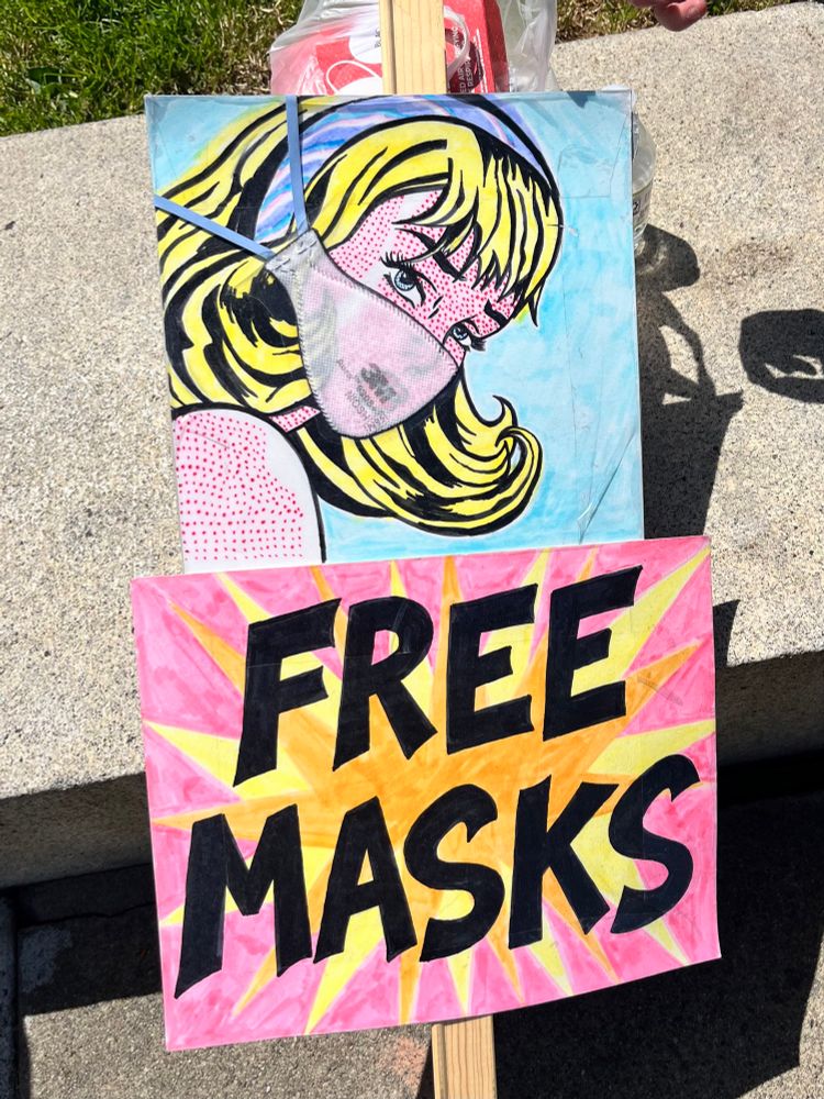 A colorful Roy Lichtenstein-style sign of a white woman with a blonde flip wearing an N95 mask and the words “free masks” in black letters on a lower sign that has a pink and orange starburst design. 
