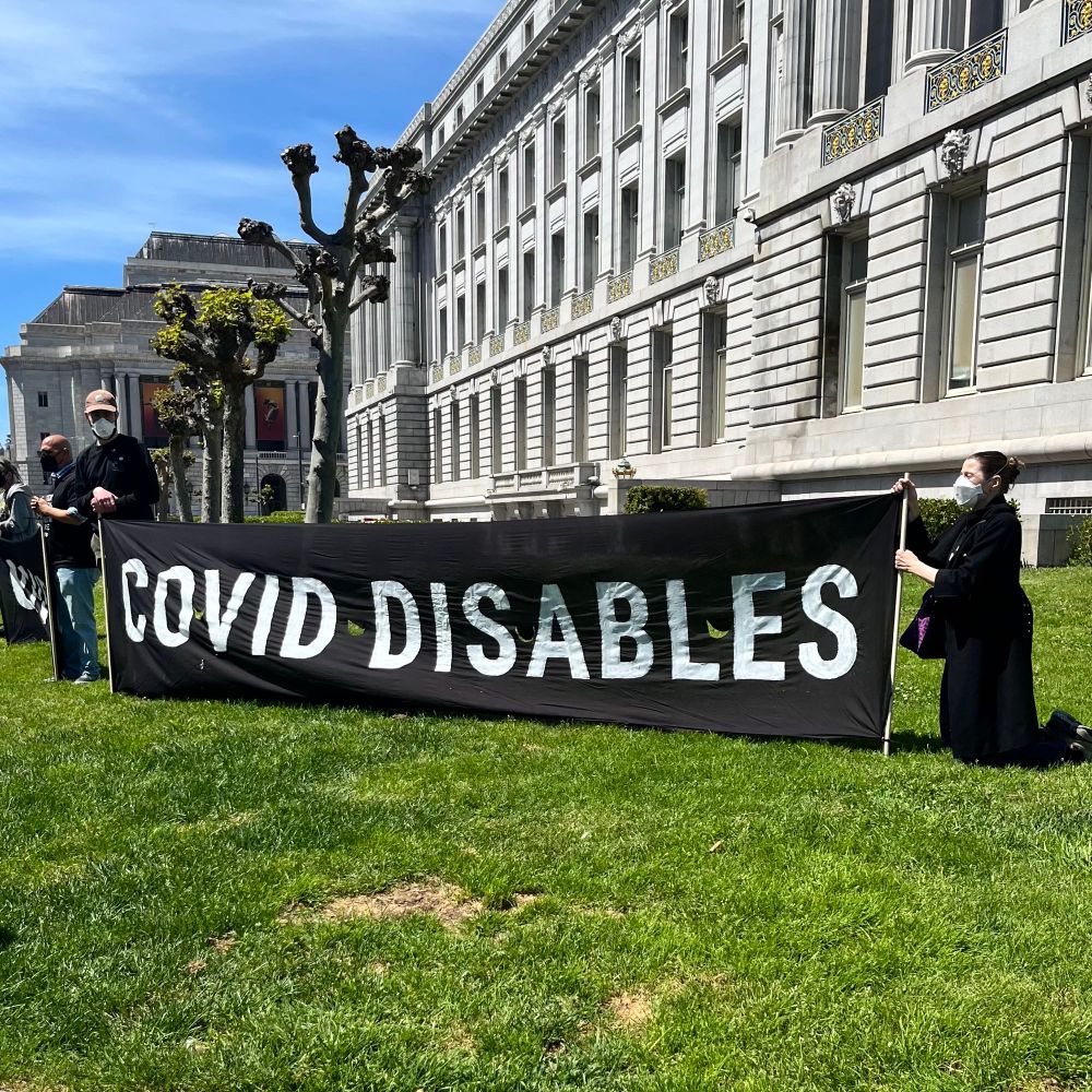 A large black and white banner that reads “Covid disables” in capital letters on a green lawn in front of a stone government building. The banner is held by two people wearing N95 masks.