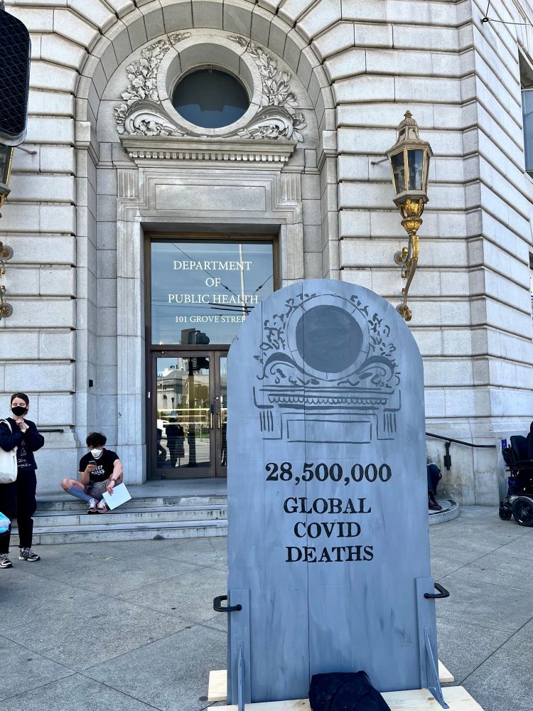 A foam tombstone in the shape of the San Francisco department of public health doorway with the words “28,500,000 global Covid deaths.” There are several people in KN95 and N95 masks standing and sitting in the background. 