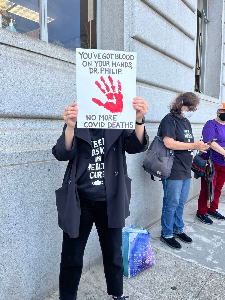 A Gran Fury-inspired bloody handprint sign that reads “You’ve got blood on your hands Dr. Philip — no more Covid deaths.” The person holding the sign covering their face is wearing black clothing and a t-shirt that reads “keep masks in healthcare.” Behind them are several people in masks against a stone building on a concrete sidewalk.