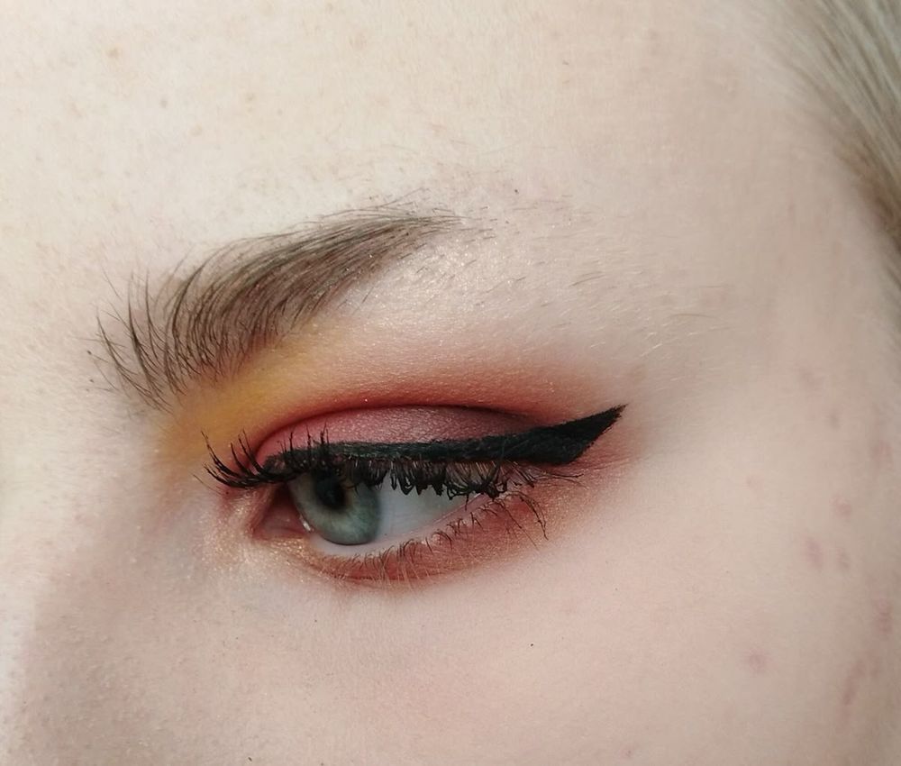 closeup photo of a person with a half-shaved eyebrow, wearing eyeliner and warm-toned eyeshadow