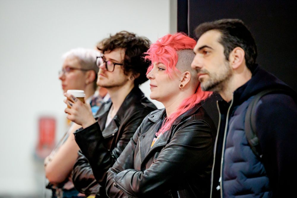 A photo of four people standing in a row, from the side. I'm the only one in focus. I have a curly pink mullet hawk, and I'm wearing a black leather jacket with my arms crossed. I look cool as fuck.
