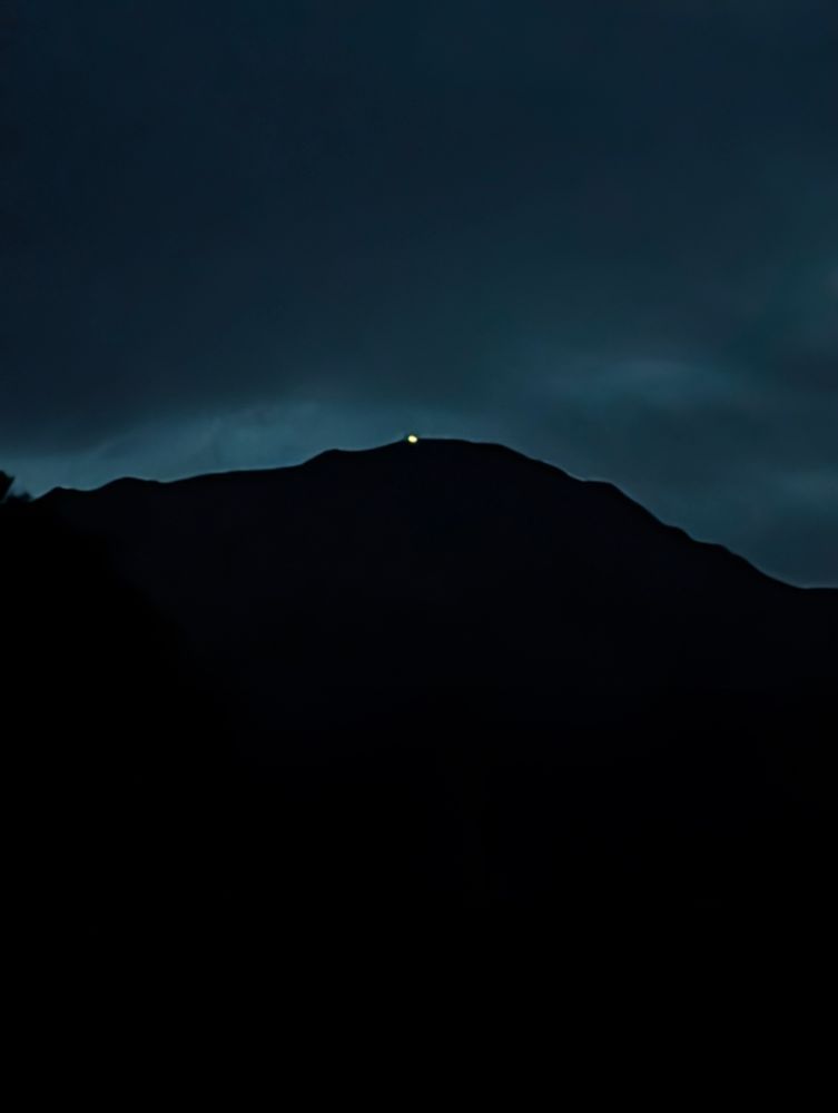 A silhouette of Pikes Peak against a dark, cloudy sky. There's a light at the summit. 