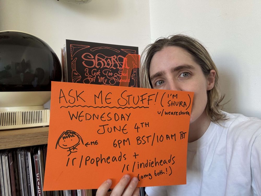 Shura holds an orange piece of paper that reads " ASK ME STUFF! (I'm Shura) Wednesday June 4th 6pm BST / 10 am PST, r/popheads + r/indieheads (omg both!)"