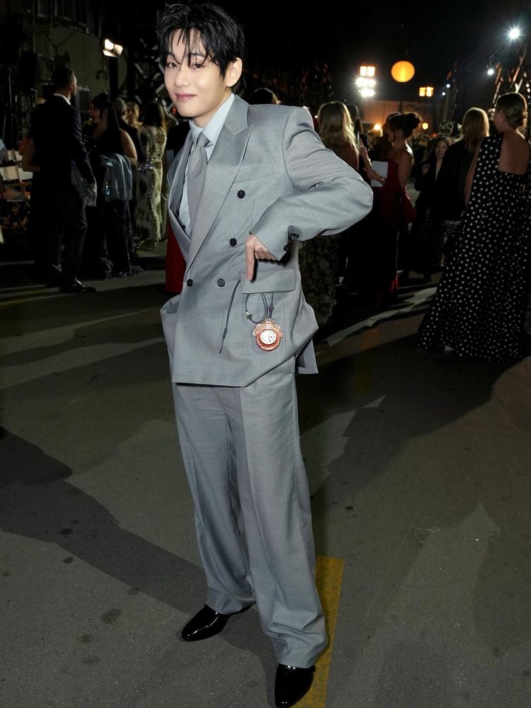 taehyung in a gray suit at the vogue hollywood event while he points to a red antique pocket watch hanging out of his pocket