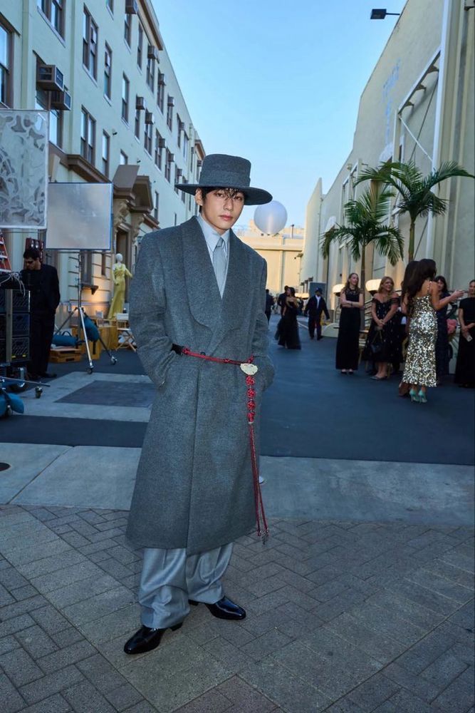taehyung in a gray suit coat with a red corded tassel featuring jade around his waist and gray hat on his head at the vogue hollywood event