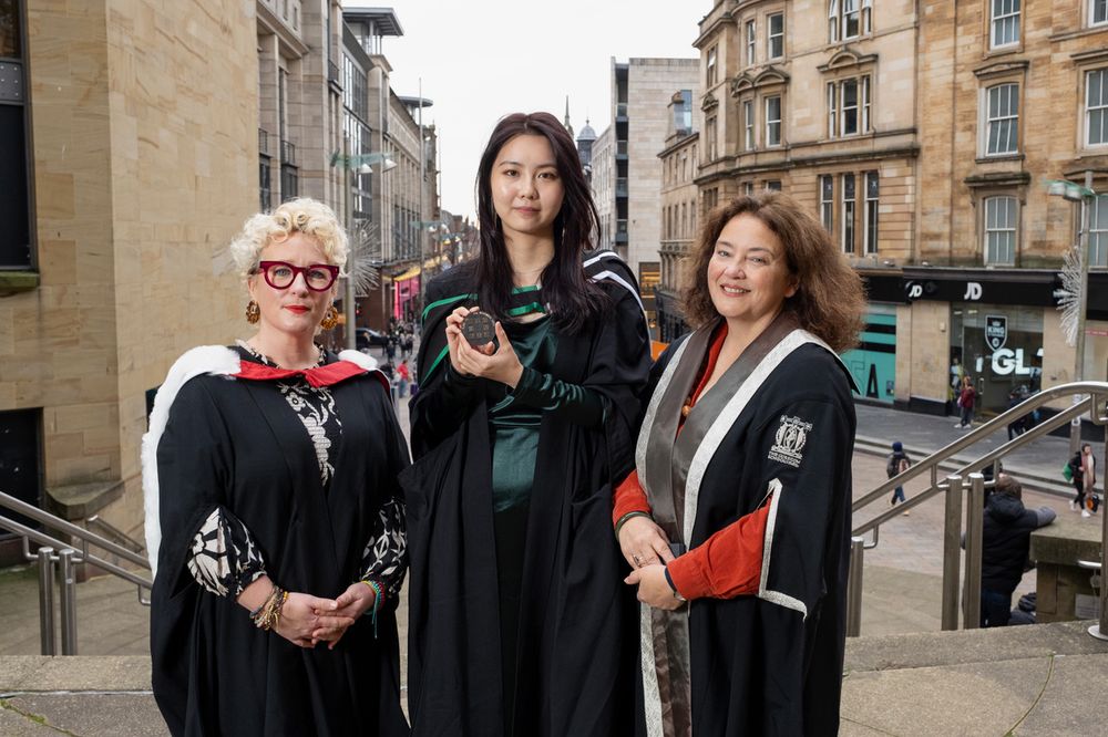 Rebecca Xu in graduation robes holds the Foulis Medal, standing between GSA Director Prof Penny MacBeth and Deputy Director (Academic) Rachel Dickson. The background is the view downhill down Glasgow's Buchanan Street, from the Royal Concert Hall