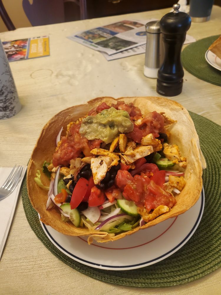 A taco salad in a baked burrito bowl: romaine, red onion, radish, seasoned chicken, tomatoes, poblano pepper, mini cucumbers, topped with salsa and guacamole. Bright colors, lovely presentation. 