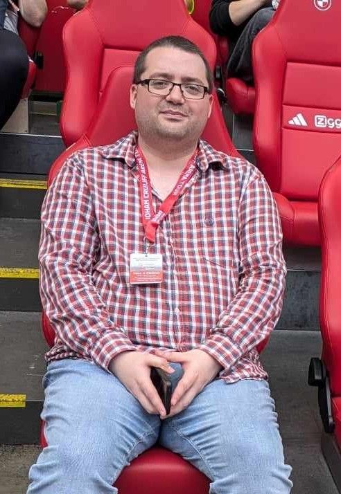 A picture of me, wearing a chequered shirt and jeans, on a leather seat inside a stadium.