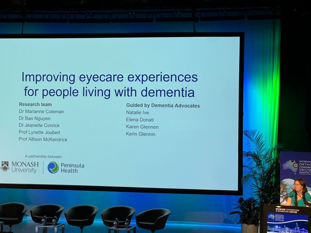 I stand at the podium in a green top next to a large screen with a slide saying "Improving eyecare experiences for people living with dementia".