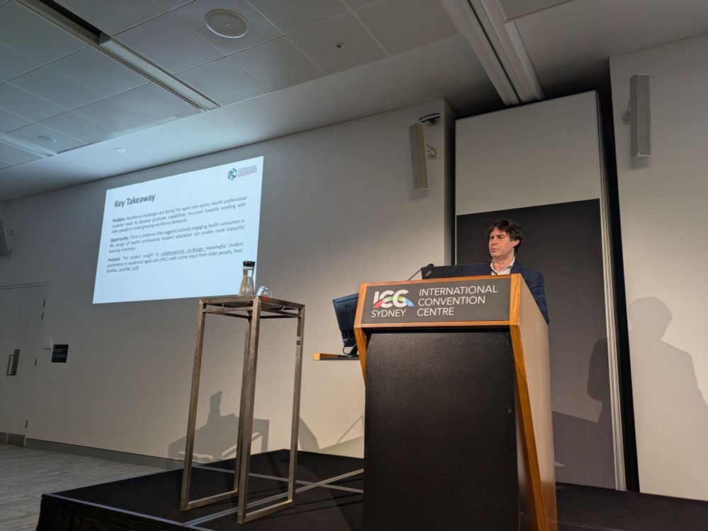 Simon in a navy suit stands at the podium with slides