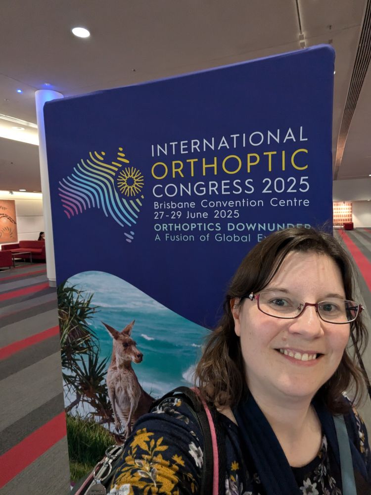 Selfie of me in a floral top and red glasses next to a banner saying International Orthoptic Congress 2025