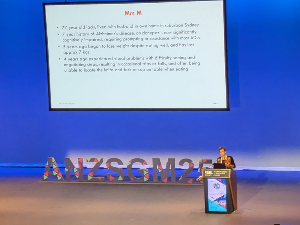 Sue in a patterned top stands at a podium with slides on a large stage in a theatre full of people