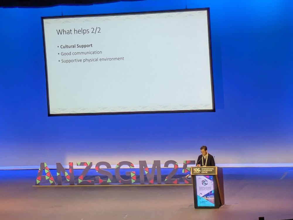 Oliver in a dark suit stands at the podium on a large stage with slides in a full theatre