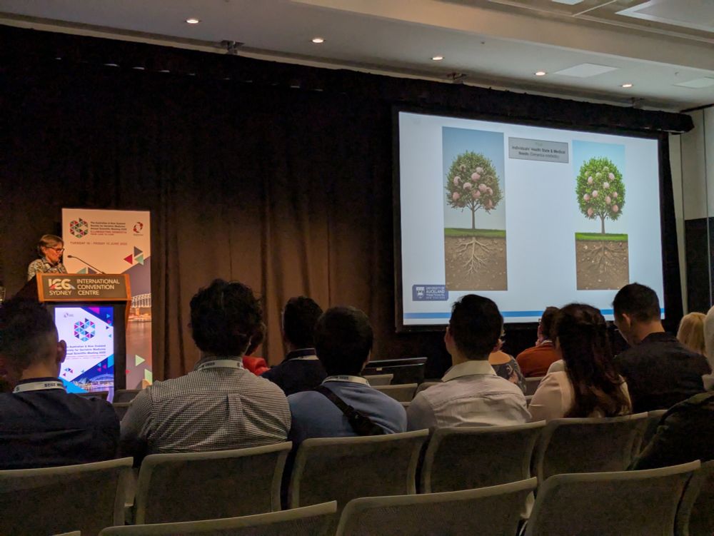 Ngaire in a patterned top stands at a podium next to slides showing a Withered tree and a blooming tree