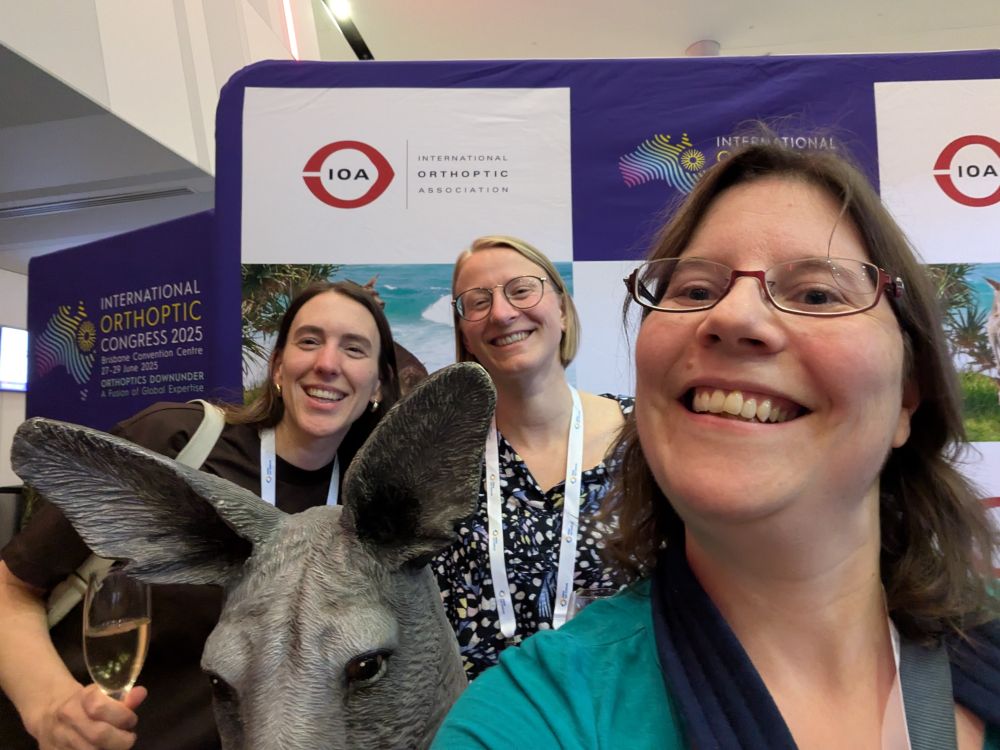 Selfie with myself in a green top and red glasses with brown hair, and two other women (brown hair, silver earrings and a dark top, blonde hair, wire glasses and black and white patterned top). We're all smiling at the camera and there is a grey kangaroo statue poking his head into the image in the bottom right corner. The backdrop reads "International Orthoptic Association"