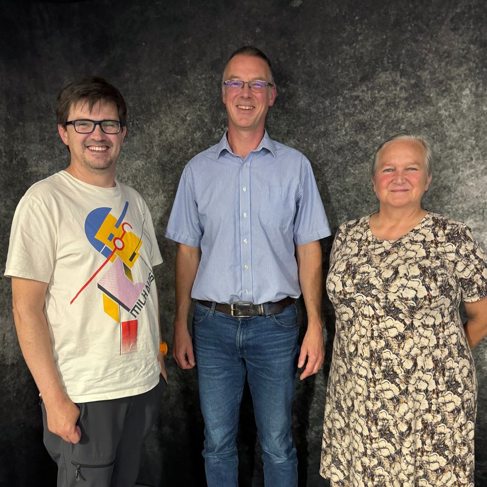 Paul Turner, Adrian Friday and Jan Bebbington smiling at the camera, standing in front of a dark grey, mottled wall.