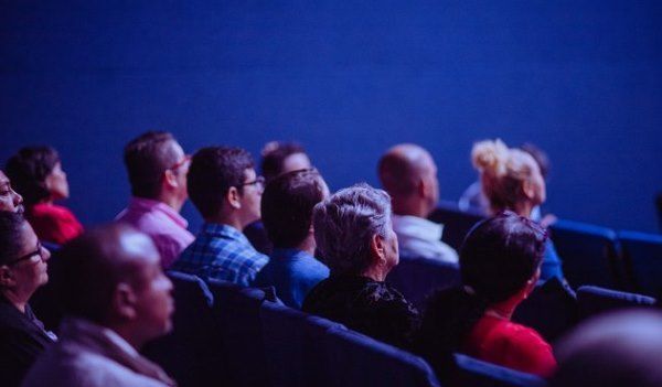 A group of people sitting in rows of seats looking forward, as if at a stage or speaker. The wall behind them is deep blue.