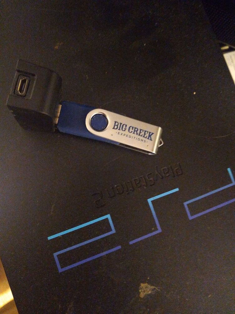 A somewhat battered USB flash drive plugged into a right angle USB type A female to USB micro b female adapter resting on top of a PlayStation 2 Fat model. The USB is branded with Big Creek Expeditions.