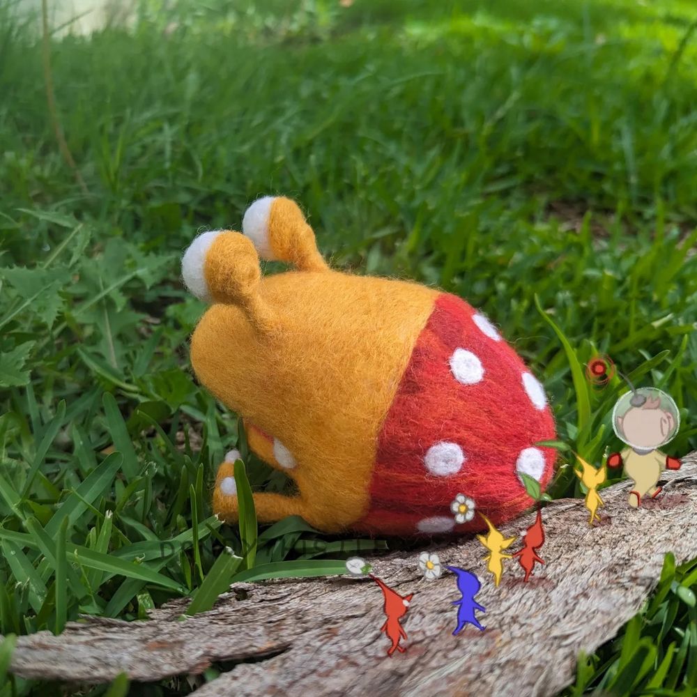 Photo of the felted bulborb from behind, mouth closed and sitting in some grass. Olimar and some Pikmin have been digitally drawn in, walking along a piece of bark past the bulborb.