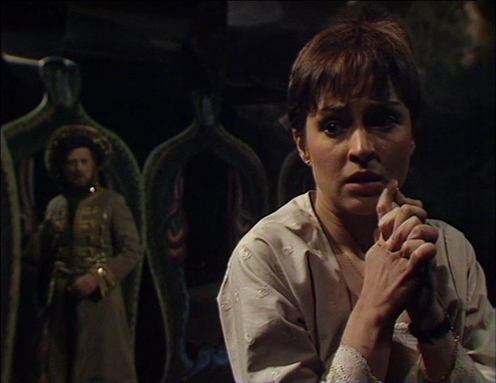 Tegan sits in the hall of mirrors. Behind her, out of focus, is the showman. She looks distressed and anxious.