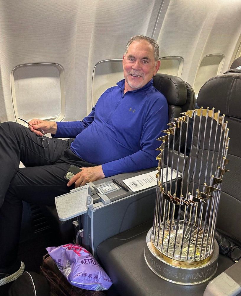 Bruce Bochy smiles proudly while sitting with one leg crossed easily over the other on an airplane. The 2023 World Series trophy is on the seat next to him. 