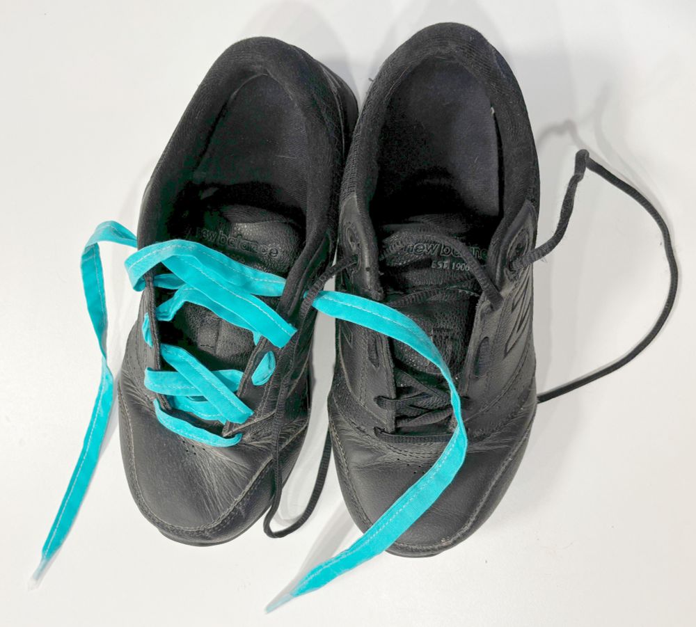 Two black sneakers. One has round black laces and the other has homemade flat blue laces.