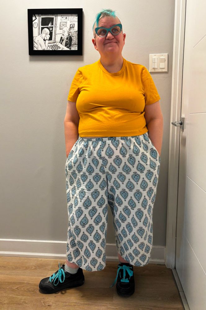 Person in hallway wearing a yellow t-shirt and blue and white patterned pants. Black sneakers with blue shoe laces. Shaved head, blue bangs, blue glasses frames. Ink drawing hung on the wall.