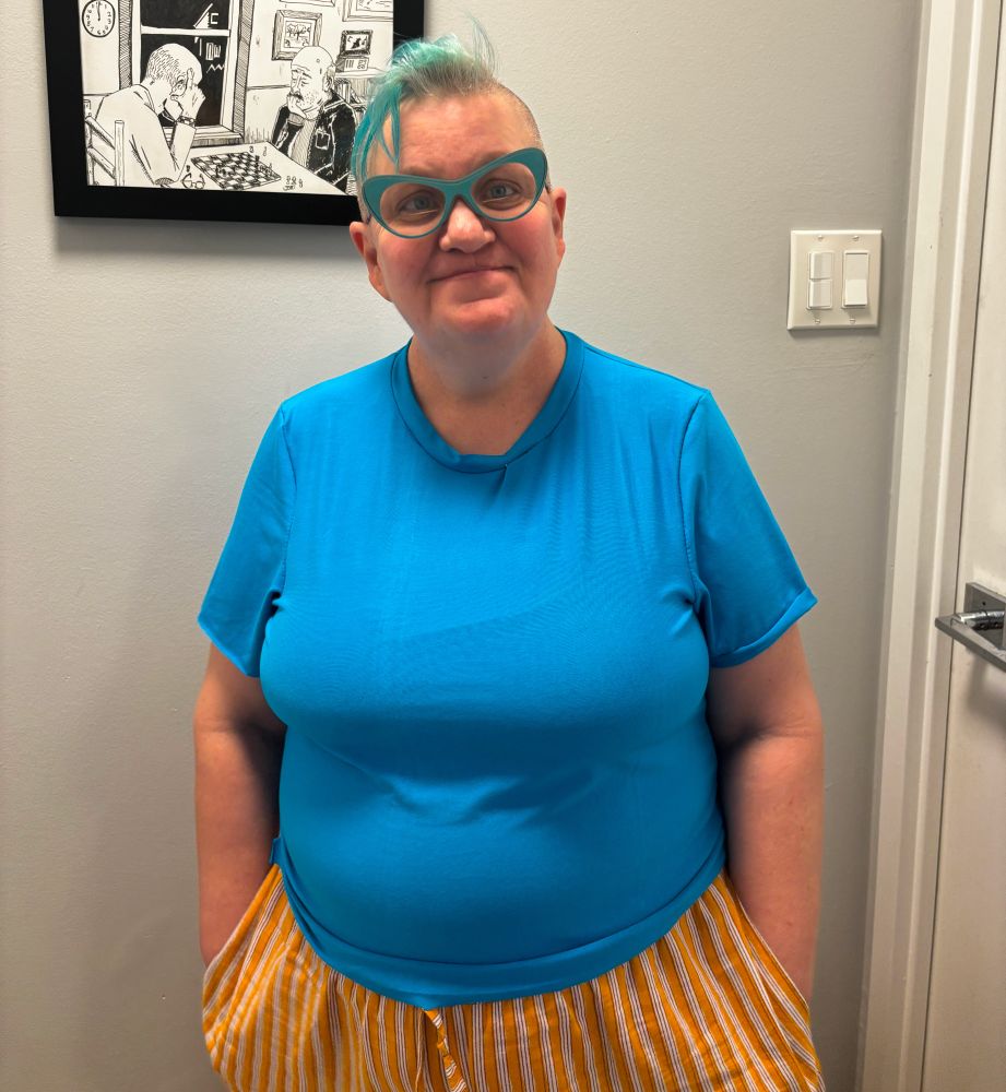 Person with blue bangs, blue glasses, and a shaved head in a blue t-shirt and yellow striped pants. Standing in a hallway in front of an ink drawing.