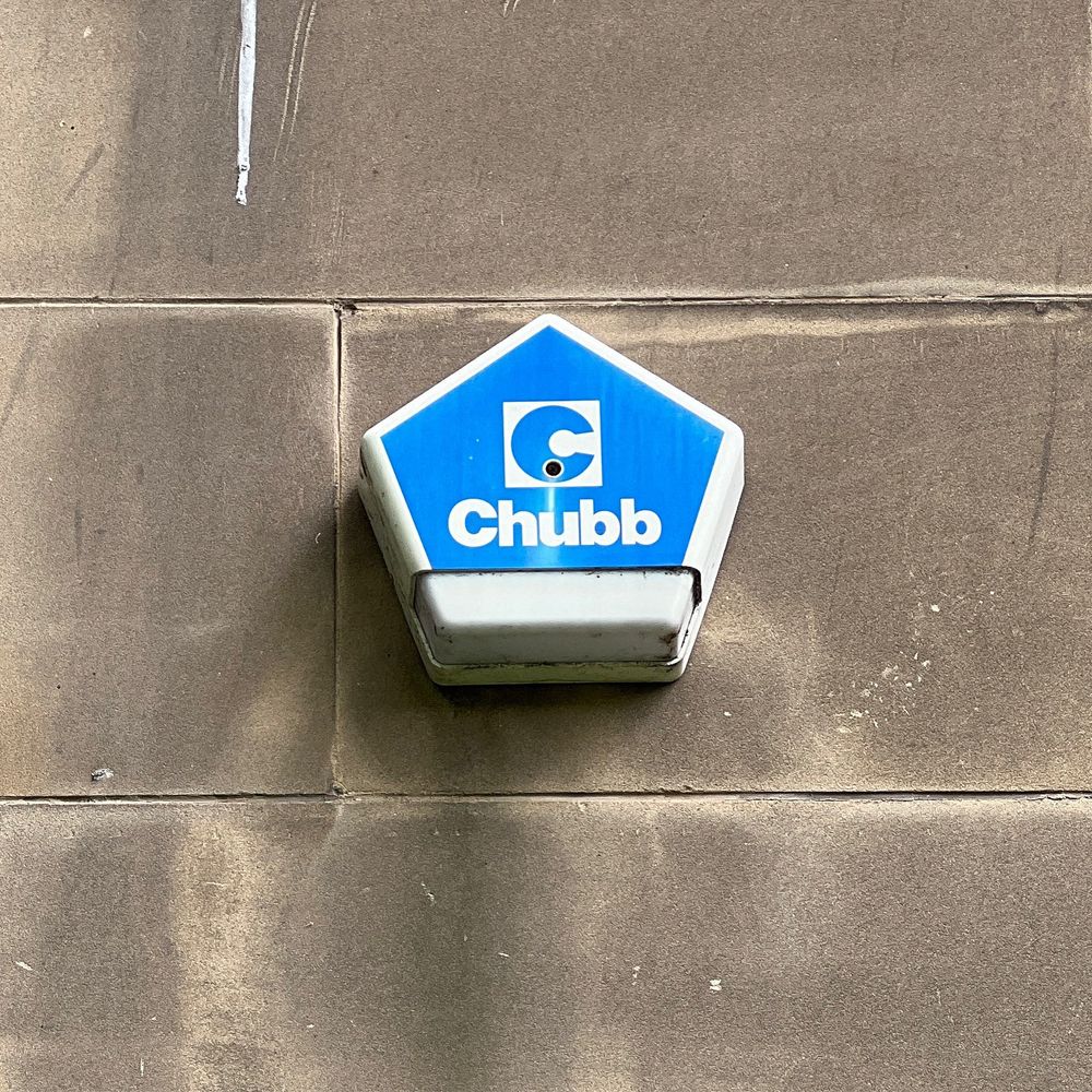 Photograph of an alarm system box on the side of a house. You can see a close up of the box on a wall. The box is pentagon shaped. 