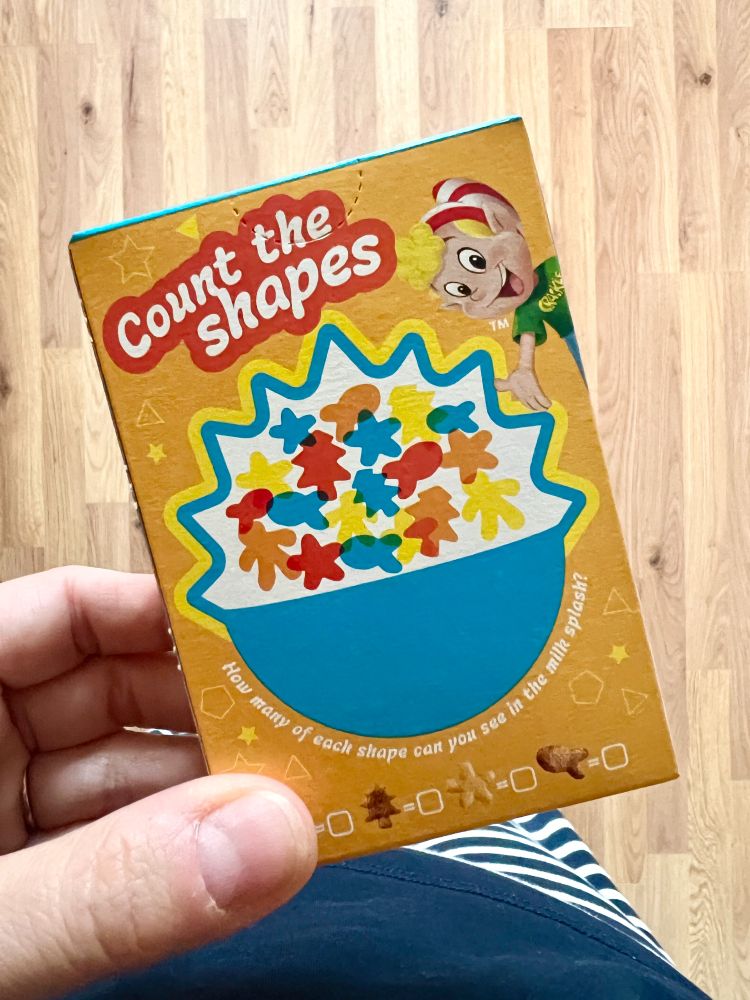 Photograph of a hand holding a small cereal packet, which has a shape counting activity on the back of it. The packet says Count the Shapes and shows an illustration of a bowl filled with cereal shapes such as stars, fish and trees.